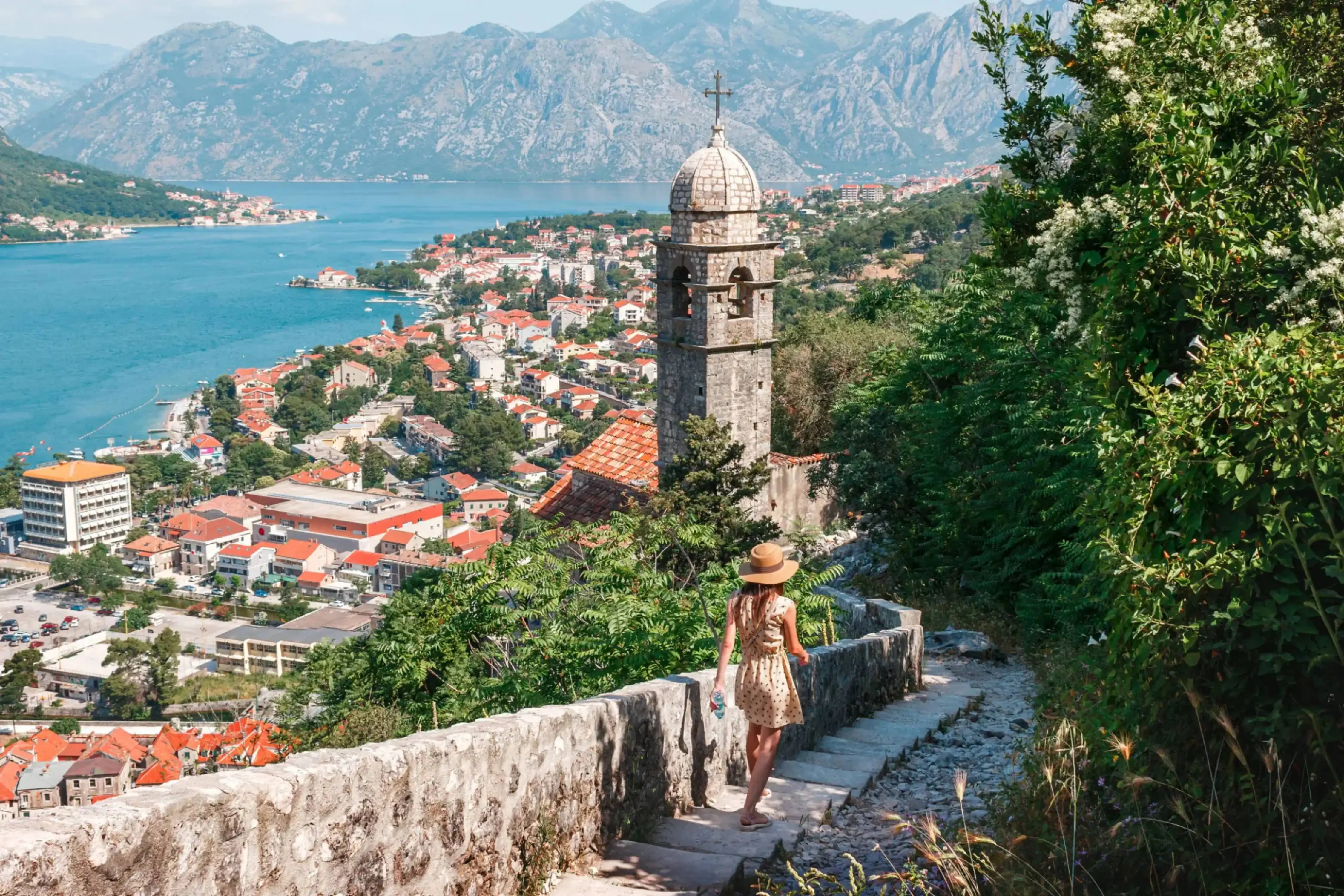 Woman walking down a stone path toward the Bay of Kotor, with a historic bell tower and red-roofed town nestled along Montenegro's Adriatic coastline.