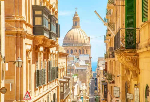 Narrow street view in Valletta, Malta, showcasing sandstone buildings, traditional Maltese balconies, and the dome of the Basilica of Our Lady of Mount Carmel in the distance.