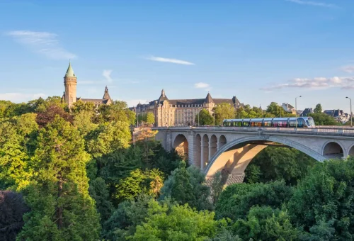 Panoramic view of the Adolphe Bridge in Luxembourg City, Luxembourg, with a colorful train crossing and the historic Banque et Caisse d'Épargne de l'État building in the background amidst lush greenery.