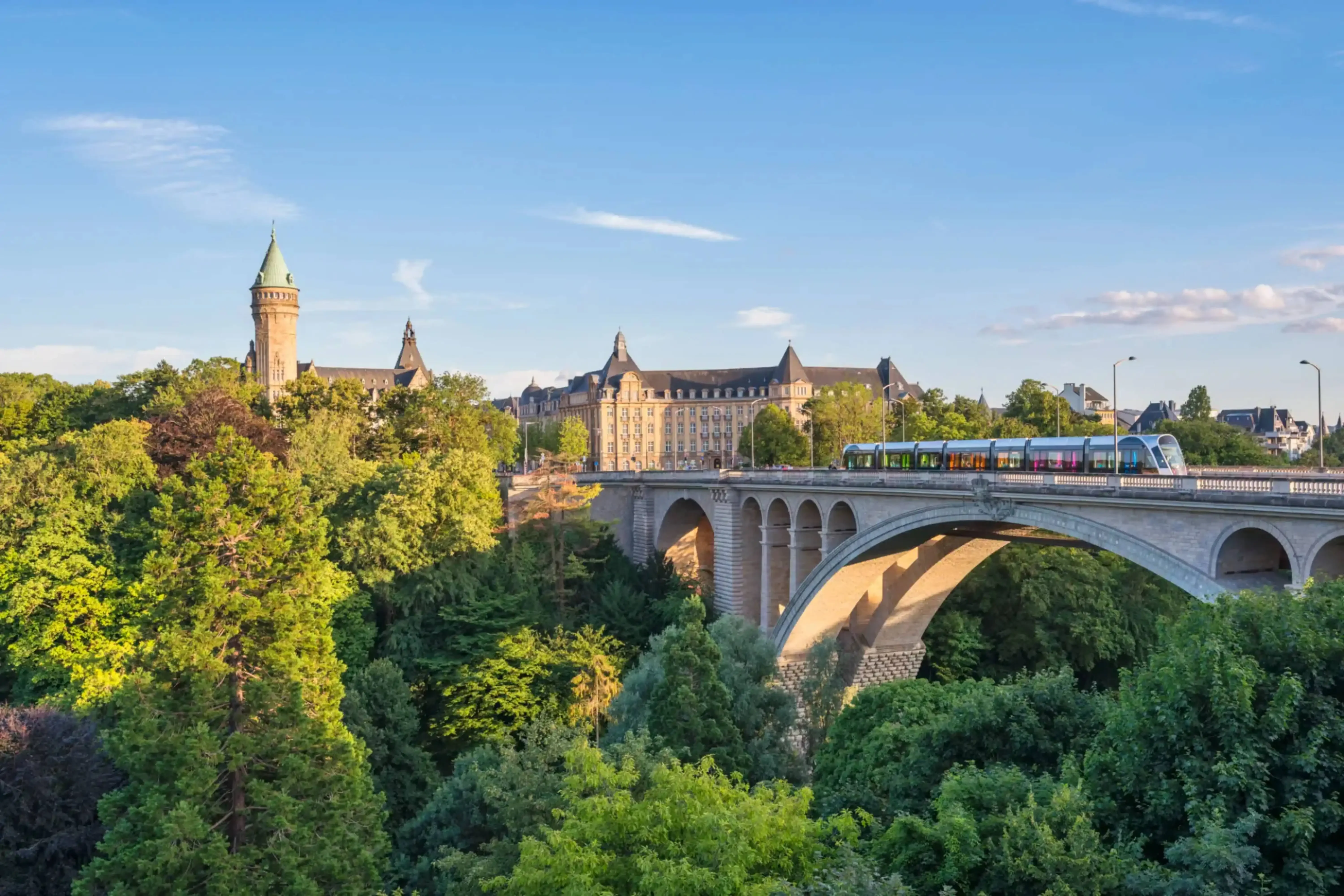 Panoramic view of the Adolphe Bridge in Luxembourg City, Luxembourg, with a colorful train crossing and the historic Banque et Caisse d'Épargne de l'État building in the background amidst lush greenery.