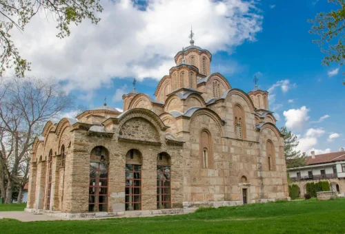 Medieval stone Orthodox monastery with multiple domes and arched windows, set on a green lawn under a partly cloudy sky.