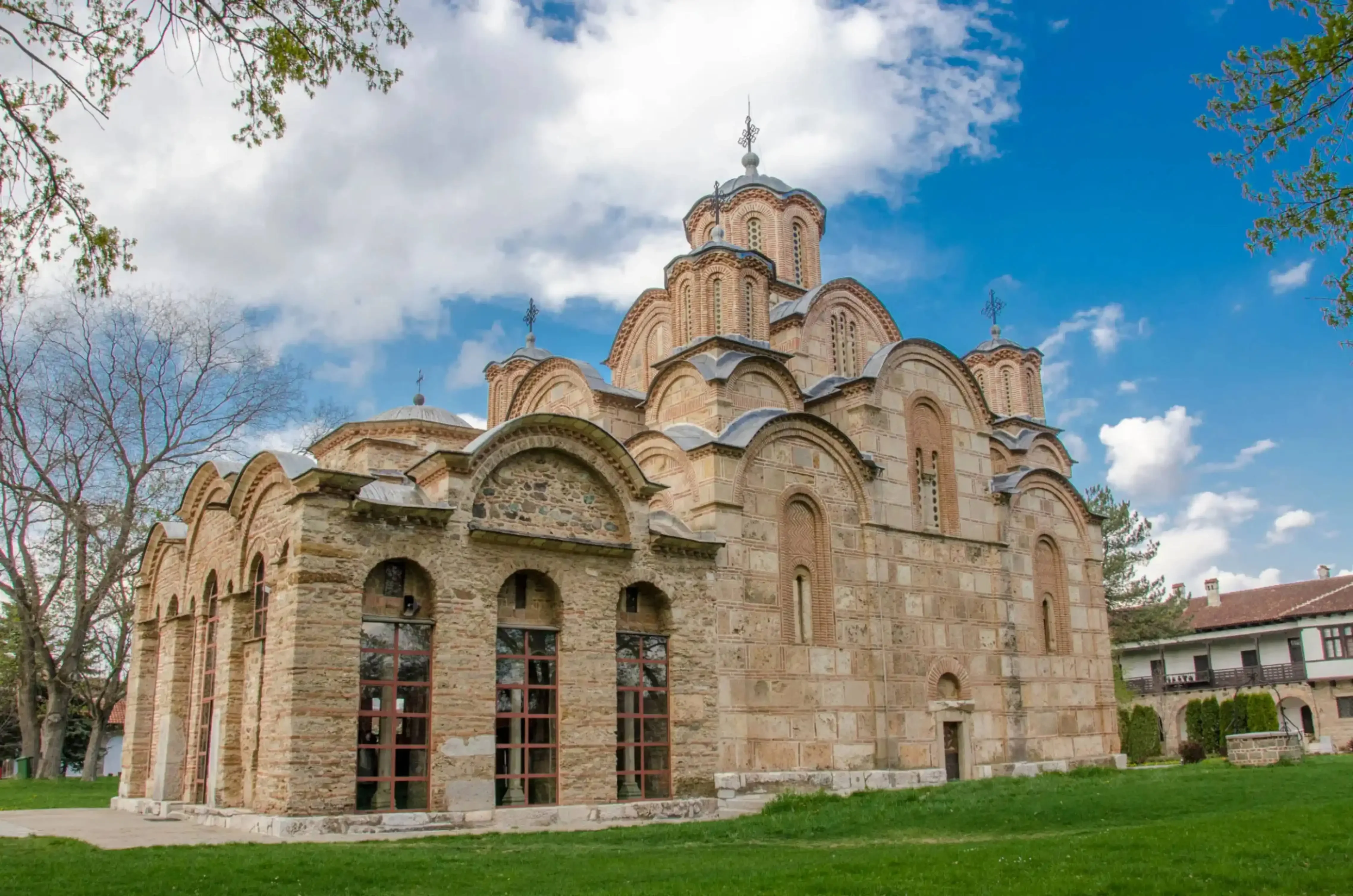 Medieval stone Orthodox monastery with multiple domes and arched windows, set on a green lawn under a partly cloudy sky.