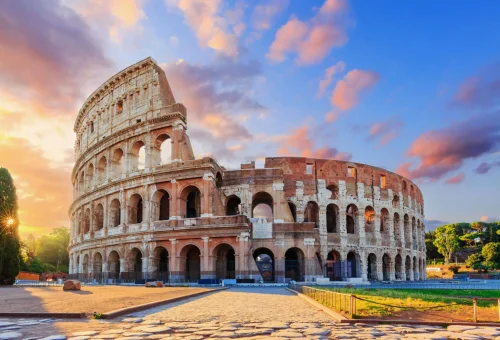 Iconic Roman Colosseum bathed in warm sunlight, with a dramatic colorful sky in the background and cobblestone ground in the foreground.