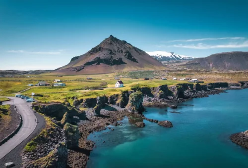 Scenic coastal road with dramatic sea cliffs and a cone-shaped volcanic mountain in the background, under a clear blue sky in Iceland.