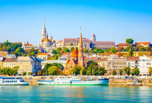 Panoramic view of Budapest’s historic skyline from across the Danube River, featuring colorful buildings and iconic churches under a bright blue sky.