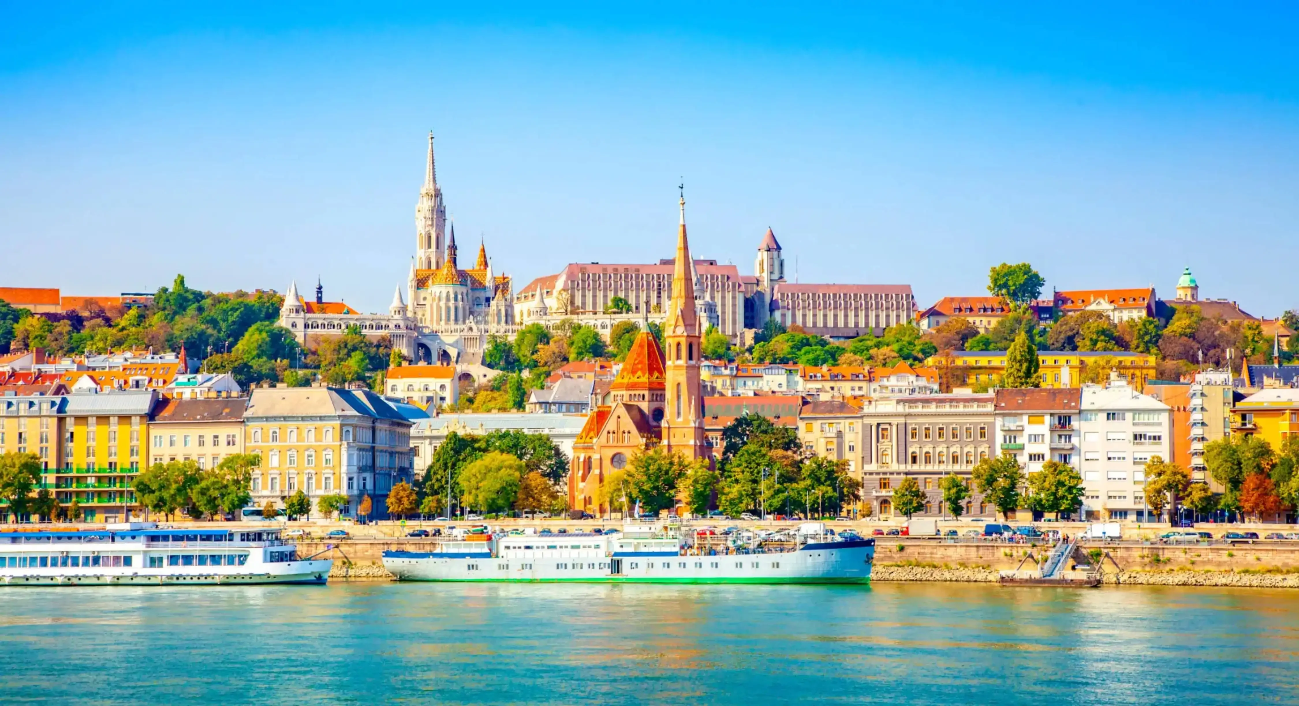 Panoramic view of Budapest’s historic skyline from across the Danube River, featuring colorful buildings and iconic churches under a bright blue sky.