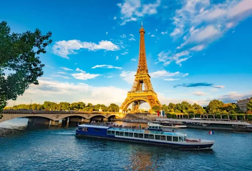 Eiffel Tower viewed from the Seine River with a sightseeing boat passing by, under a vibrant blue sky with scattered clouds in Paris, France.
