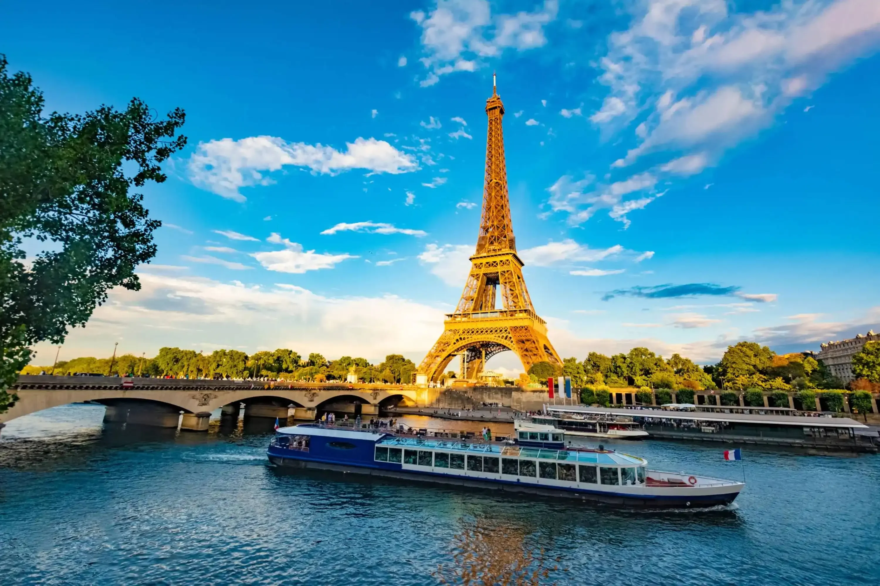 Eiffel Tower viewed from the Seine River with a sightseeing boat passing by, under a vibrant blue sky with scattered clouds in Paris, France.