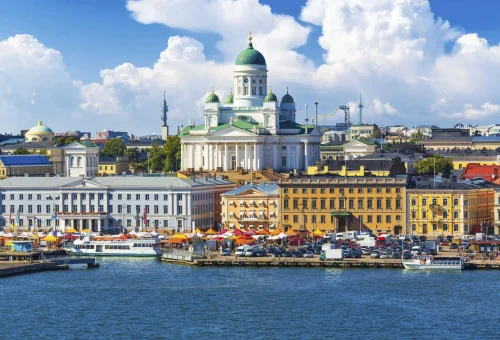 View of Helsinki Cathedral towering above colorful historic buildings along the waterfront at Market Square in Helsinki, Finland, with vibrant umbrellas and boats in the harbor under a bright blue sky.