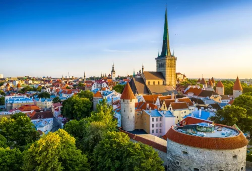 Panoramic view of Tallinn’s medieval Old Town in Estonia with red-roofed buildings, ancient stone towers, and the tall spire of St. Olaf’s Church rising prominently against a clear blue sky.