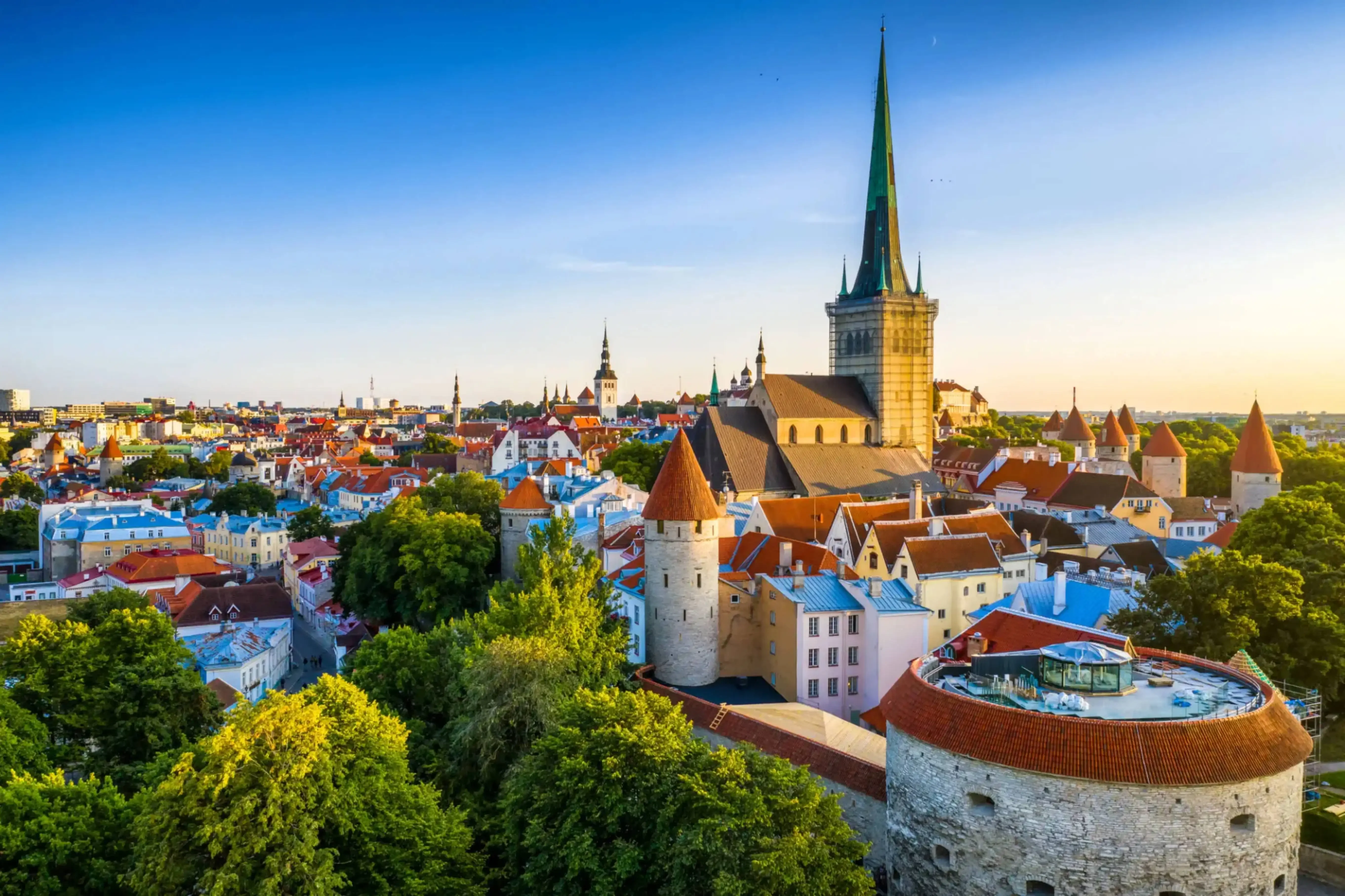 Panoramic view of Tallinn’s medieval Old Town in Estonia with red-roofed buildings, ancient stone towers, and the tall spire of St. Olaf’s Church rising prominently against a clear blue sky.