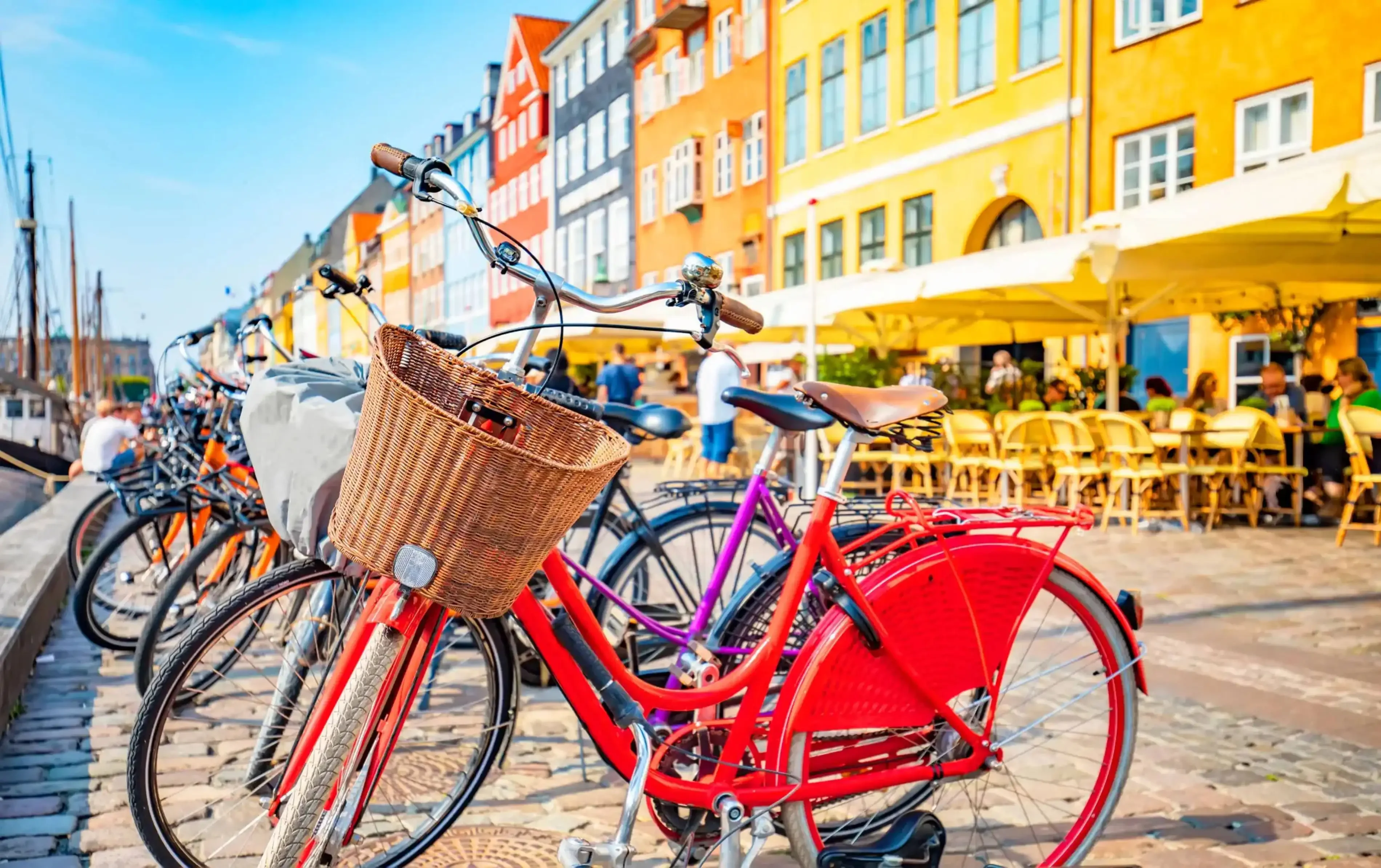 Row of colorful bicycles parked along the cobblestone harborfront of Nyhavn in Copenhagen, Denmark, with vibrant historic buildings and outdoor cafes in the background.