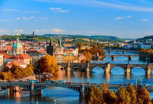 Aerial view of Prague, Czech Republic during autumn, showcasing the Vltava River with historic stone bridges, colorful trees, and the city’s skyline of spires and domes.