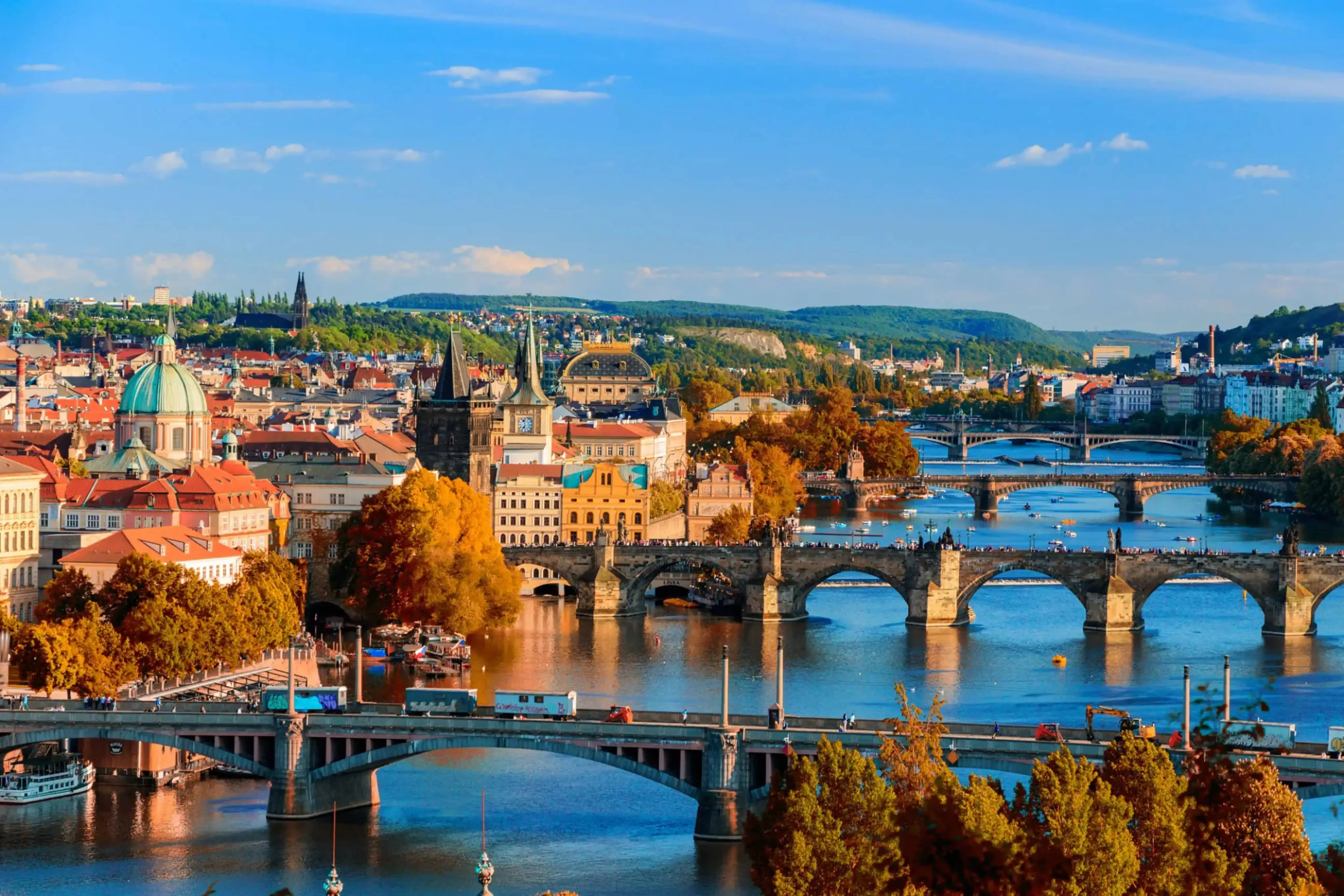Aerial view of Prague, Czech Republic during autumn, showcasing the Vltava River with historic stone bridges, colorful trees, and the city’s skyline of spires and domes.