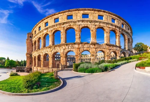 Ancient Roman amphitheater with multiple arches and weathered stone facade, surrounded by landscaped greenery and a curved modern pathway under a vivid blue sky.