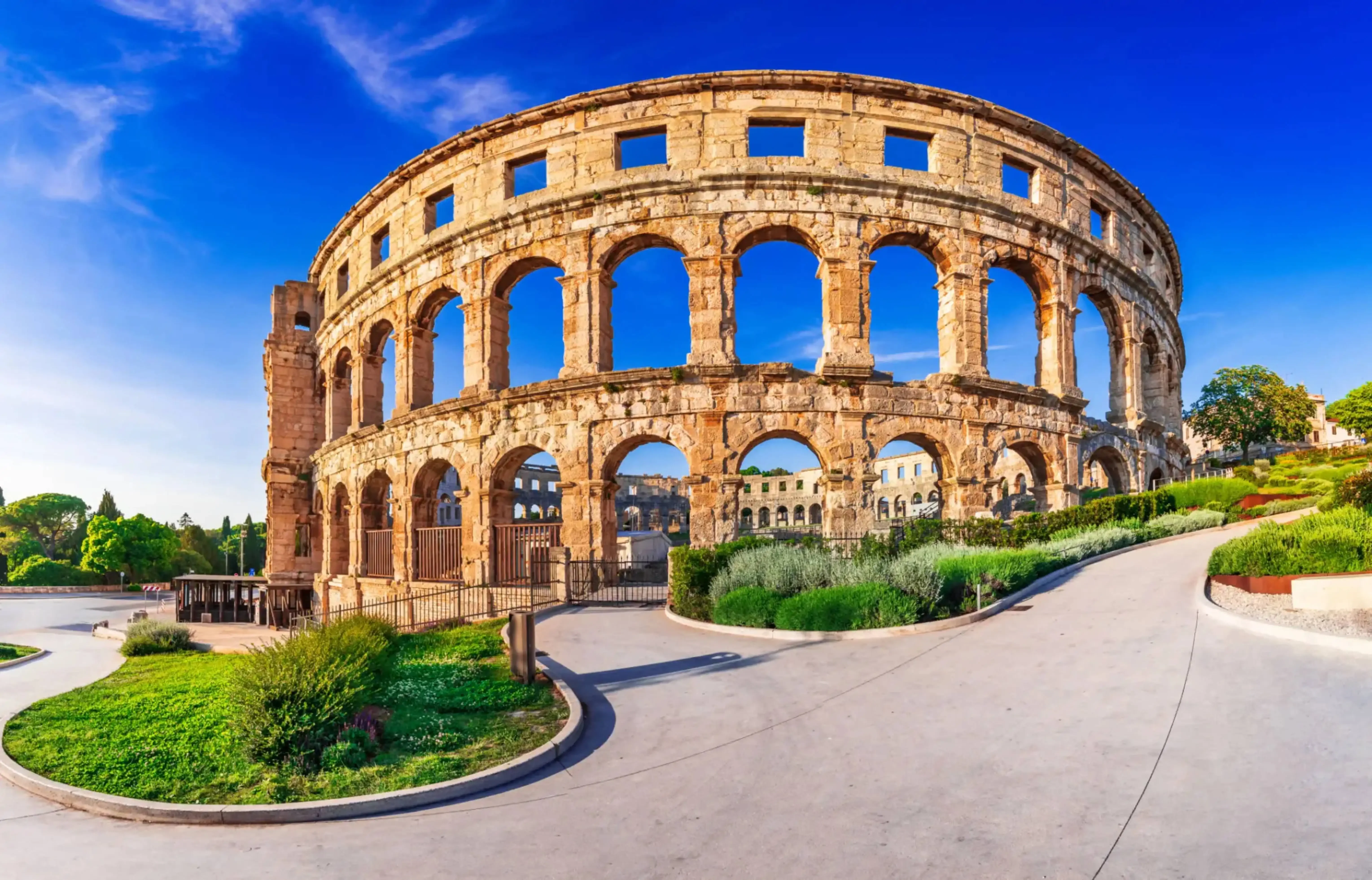 Ancient Roman amphitheater with multiple arches and weathered stone facade, surrounded by landscaped greenery and a curved modern pathway under a vivid blue sky.