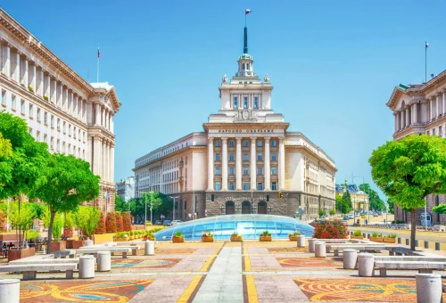 Grand neoclassical government building with a central spire, flanked by symmetrical buildings and a wide plaza with benches, patterned pavement, and manicured trees under a clear blue sky.