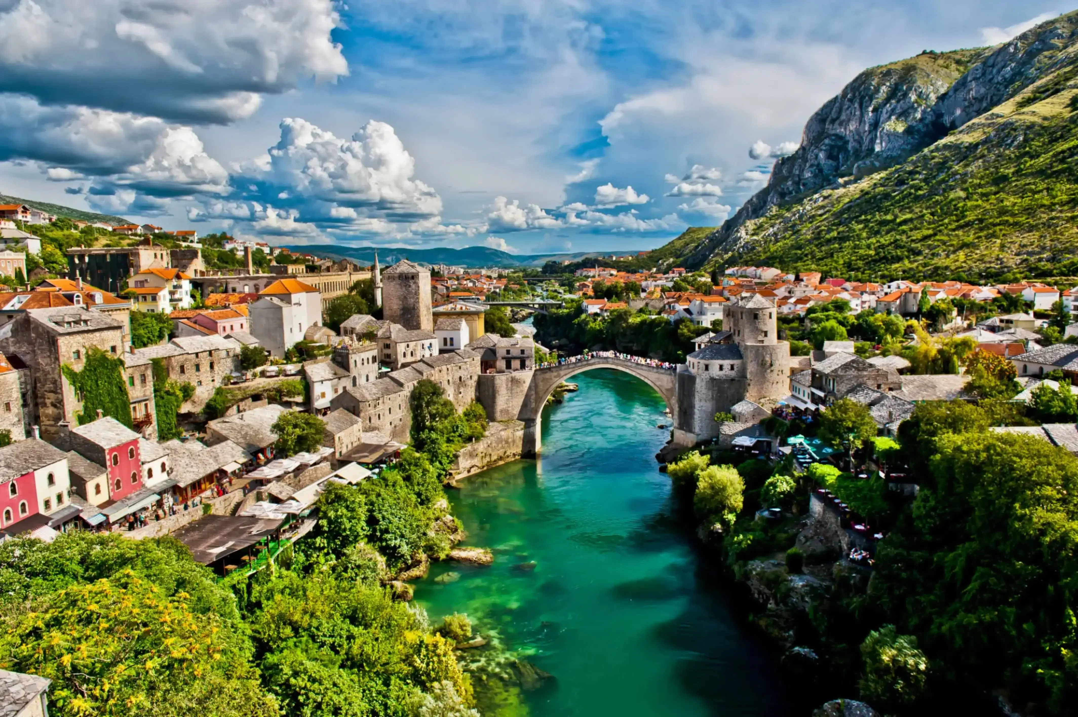 Aerial view of the historic Stari Most (Old Bridge) spanning the emerald green Neretva River, surrounded by traditional stone buildings and lush greenery in Mostar, Bosnia and Herzegovina.
