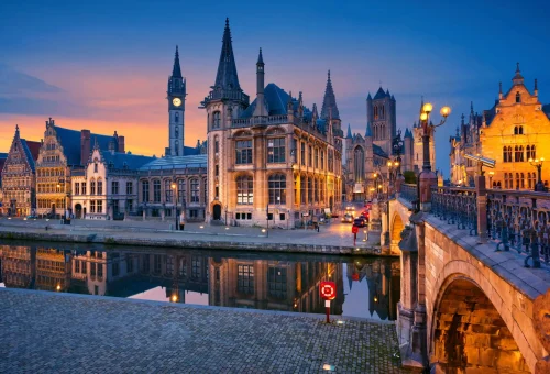 Evening view of medieval buildings and canal in Ghent, Belgium, with gothic spires and towers illuminated under a twilight sky reflecting in the calm waters.