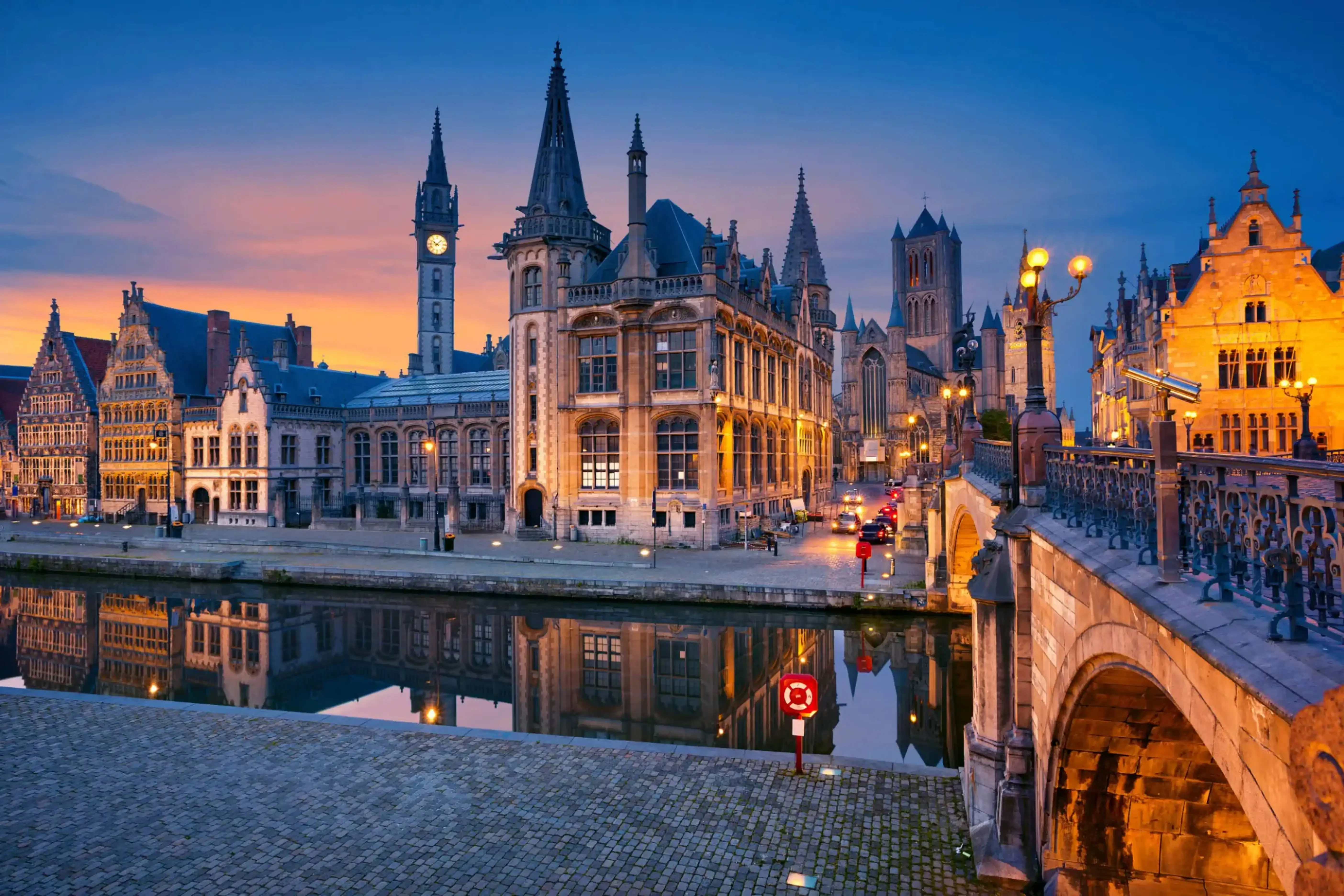Evening view of medieval buildings and canal in Ghent, Belgium, with gothic spires and towers illuminated under a twilight sky reflecting in the calm waters.