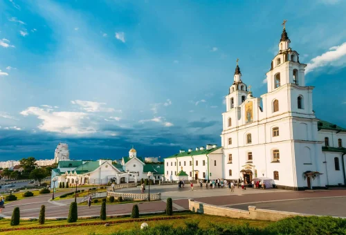 Daytime view of a historic white cathedral with twin towers and green-roofed buildings in a spacious square, with people gathered in front under a partly cloudy blue sky.