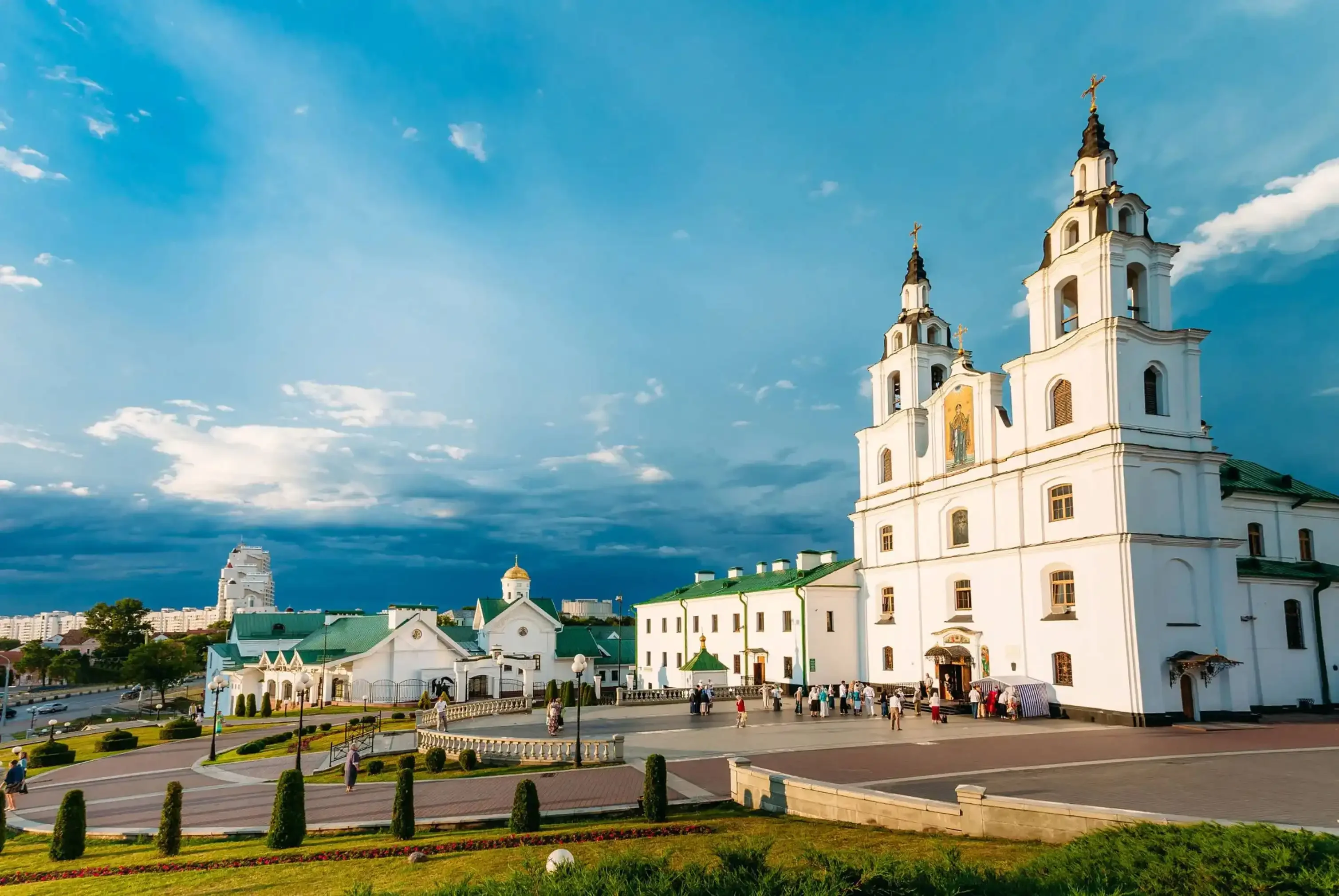 Daytime view of a historic white cathedral with twin towers and green-roofed buildings in a spacious square, with people gathered in front under a partly cloudy blue sky.