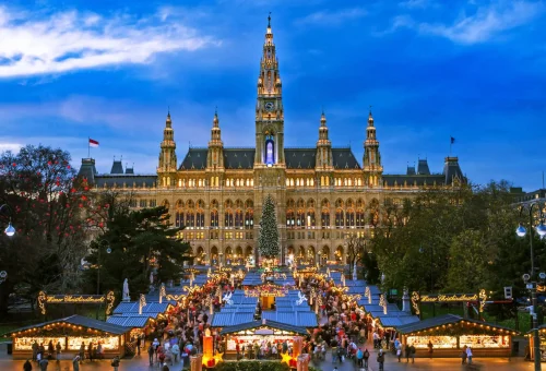Nighttime view of a grand Gothic-style building illuminated with warm lights, featuring a tall central clock tower and surrounded by a festive Christmas market with decorated stalls, a large Christmas tree, and crowds of visitors.