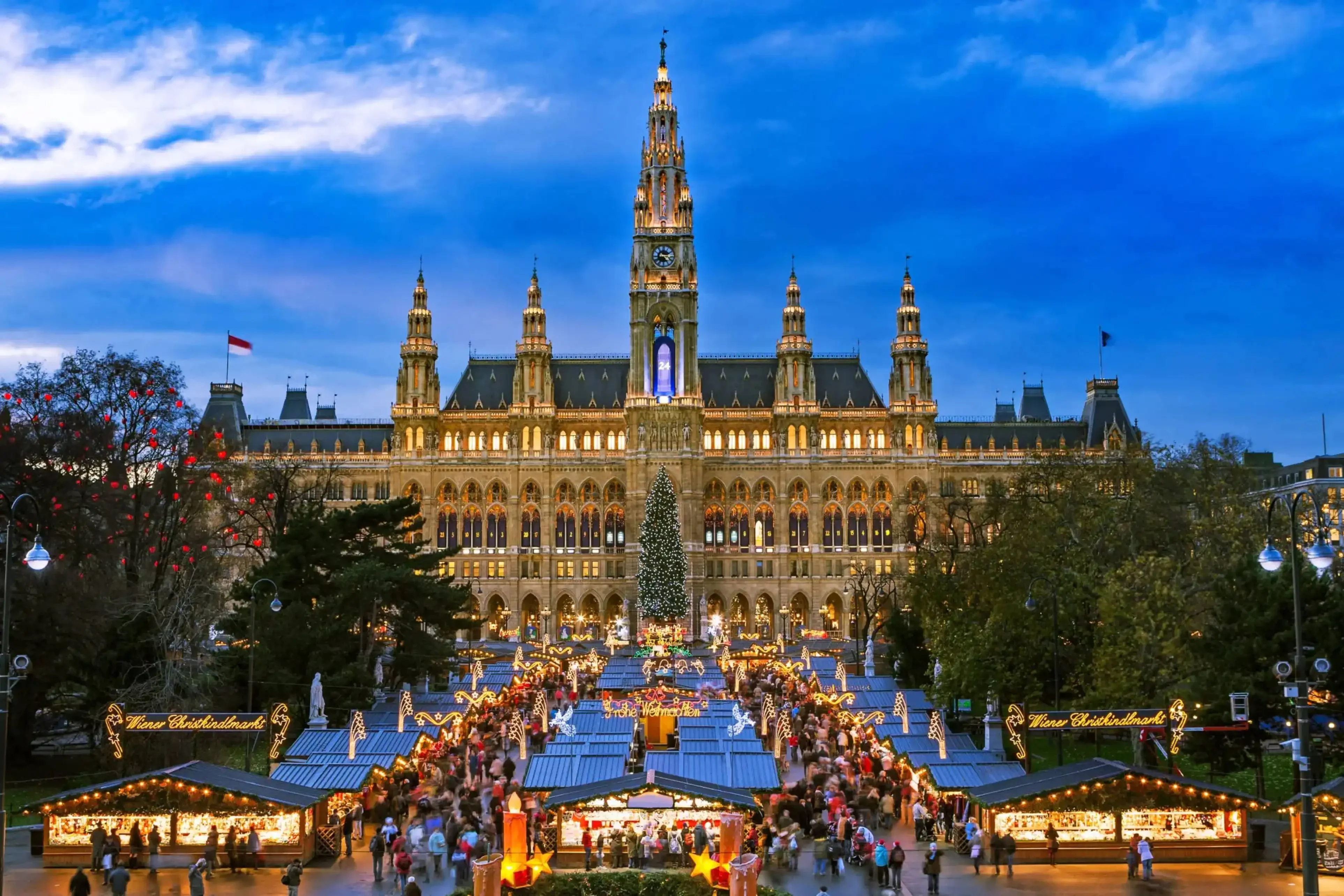 Nighttime view of a grand Gothic-style building illuminated with warm lights, featuring a tall central clock tower and surrounded by a festive Christmas market with decorated stalls, a large Christmas tree, and crowds of visitors.