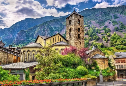 Historic stone church with a tall bell tower surrounded by vibrant flowering bushes and traditional buildings, set against a dramatic mountainous backdrop under a cloudy sky.