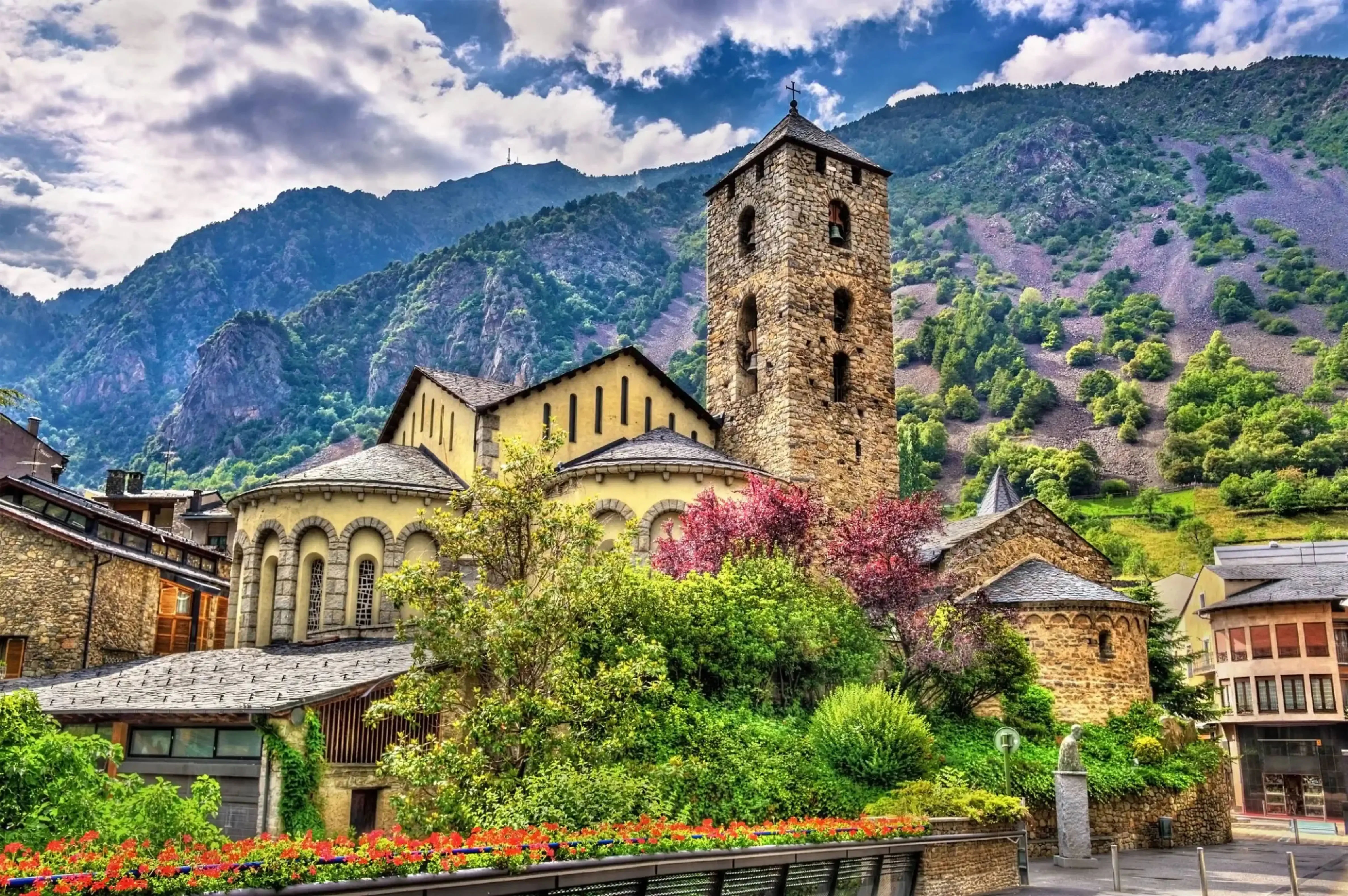 Historic stone church with a tall bell tower surrounded by vibrant flowering bushes and traditional buildings, set against a dramatic mountainous backdrop under a cloudy sky.
