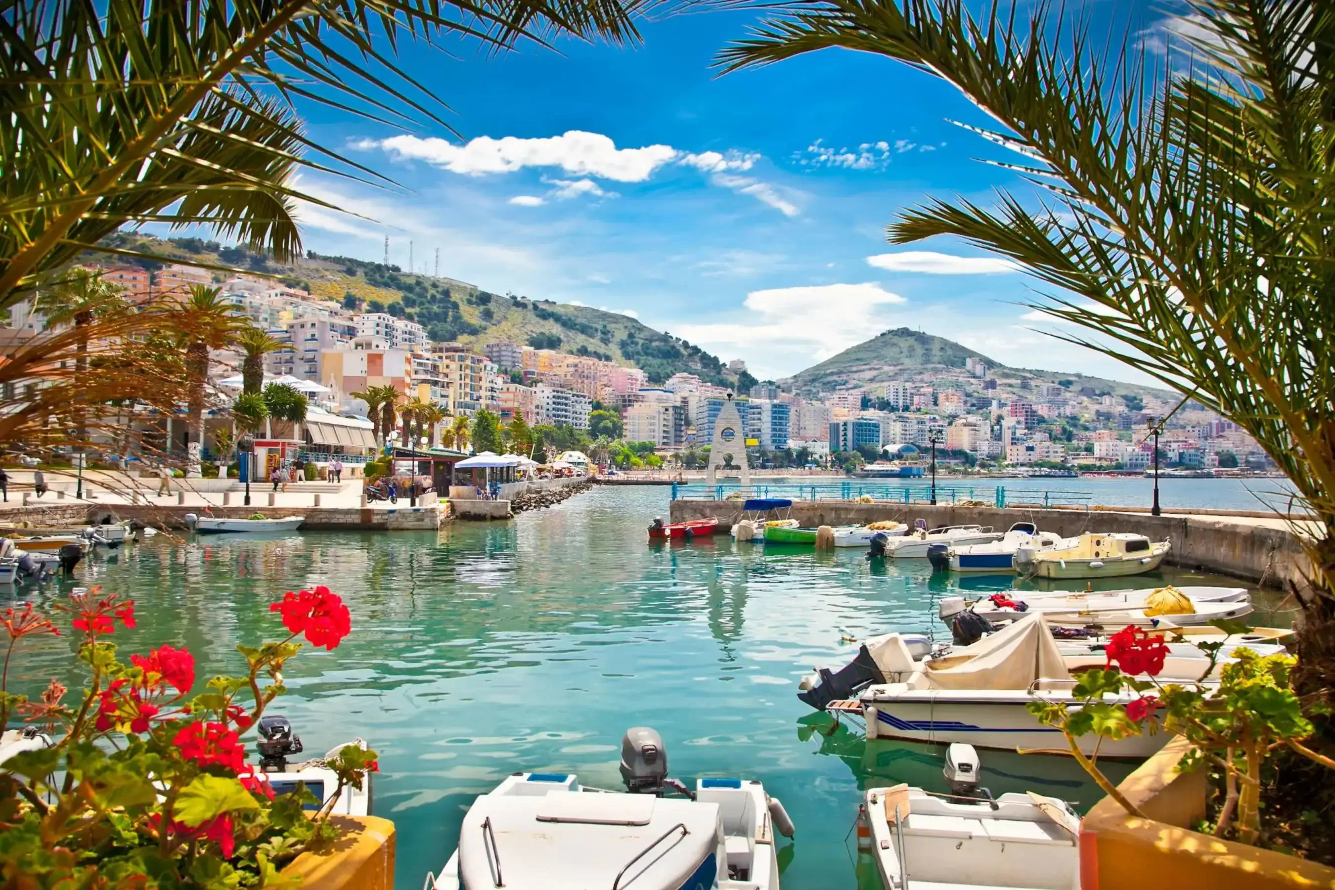 Scenic coastal harbor lined with small boats, tropical palm trees, and red flowers, with pastel-colored buildings and lush green hills in the background under a vibrant blue sky.