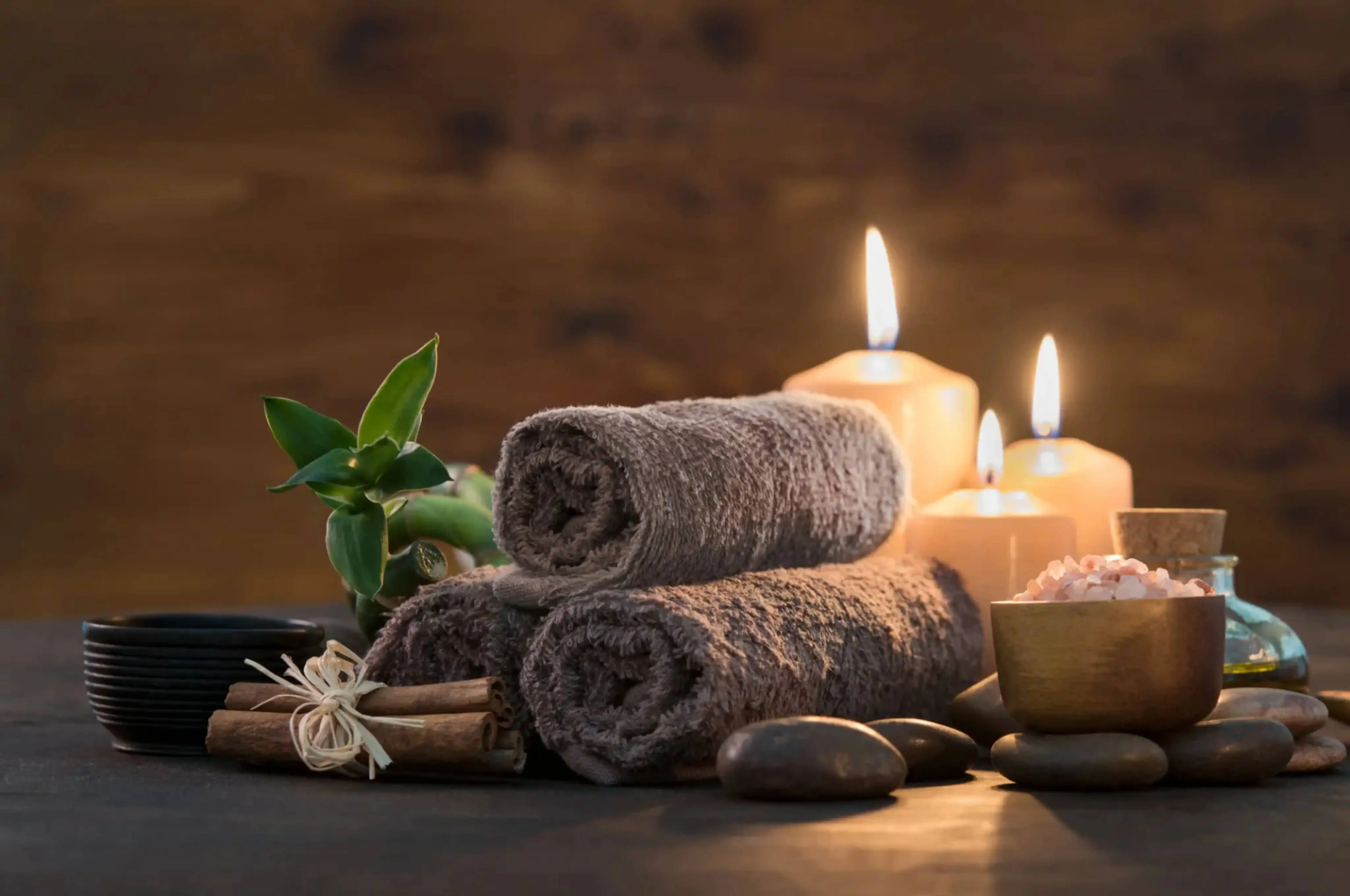 Rolled brown spa towels with candles, black massage stones, a wooden bowl of pink Himalayan salt, and green leaves arranged on a wooden surface for a relaxing wellness ambiance.