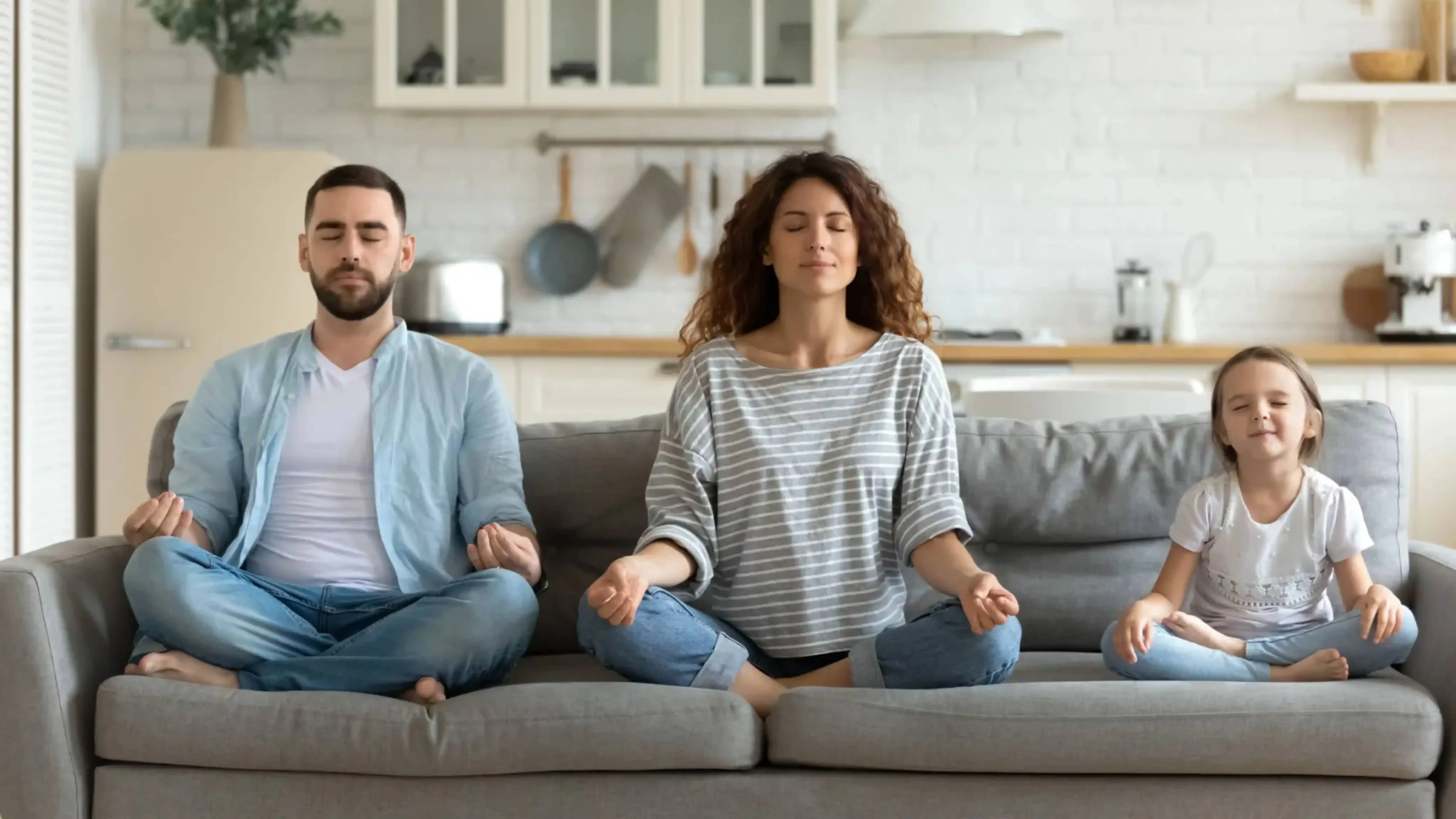 A family of three — father, mother, and young daughter — sitting cross-legged on a couch in a modern kitchen, peacefully meditating with eyes closed.