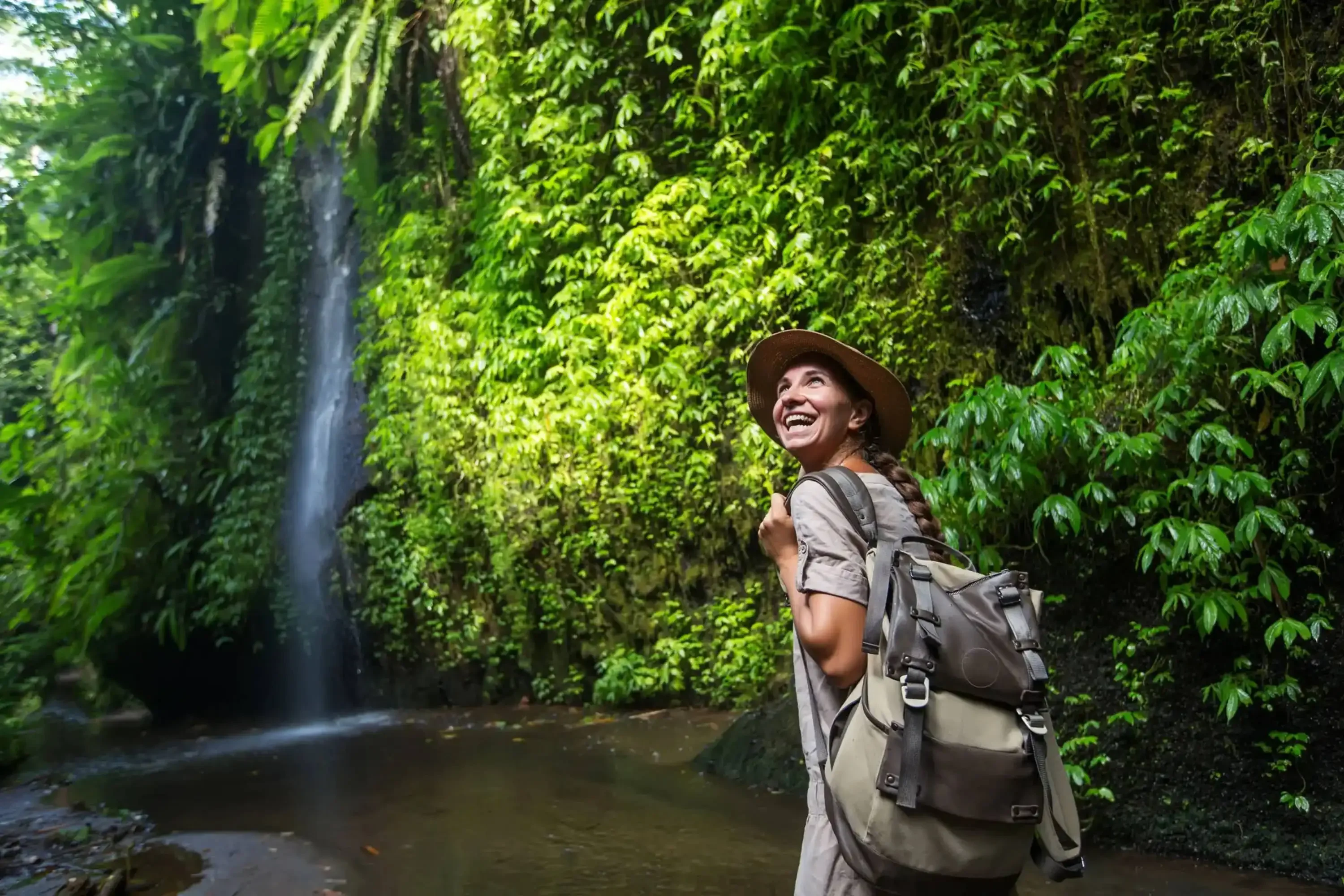 Smiling female backpacker exploring a lush rainforest trail with a waterfall in the background