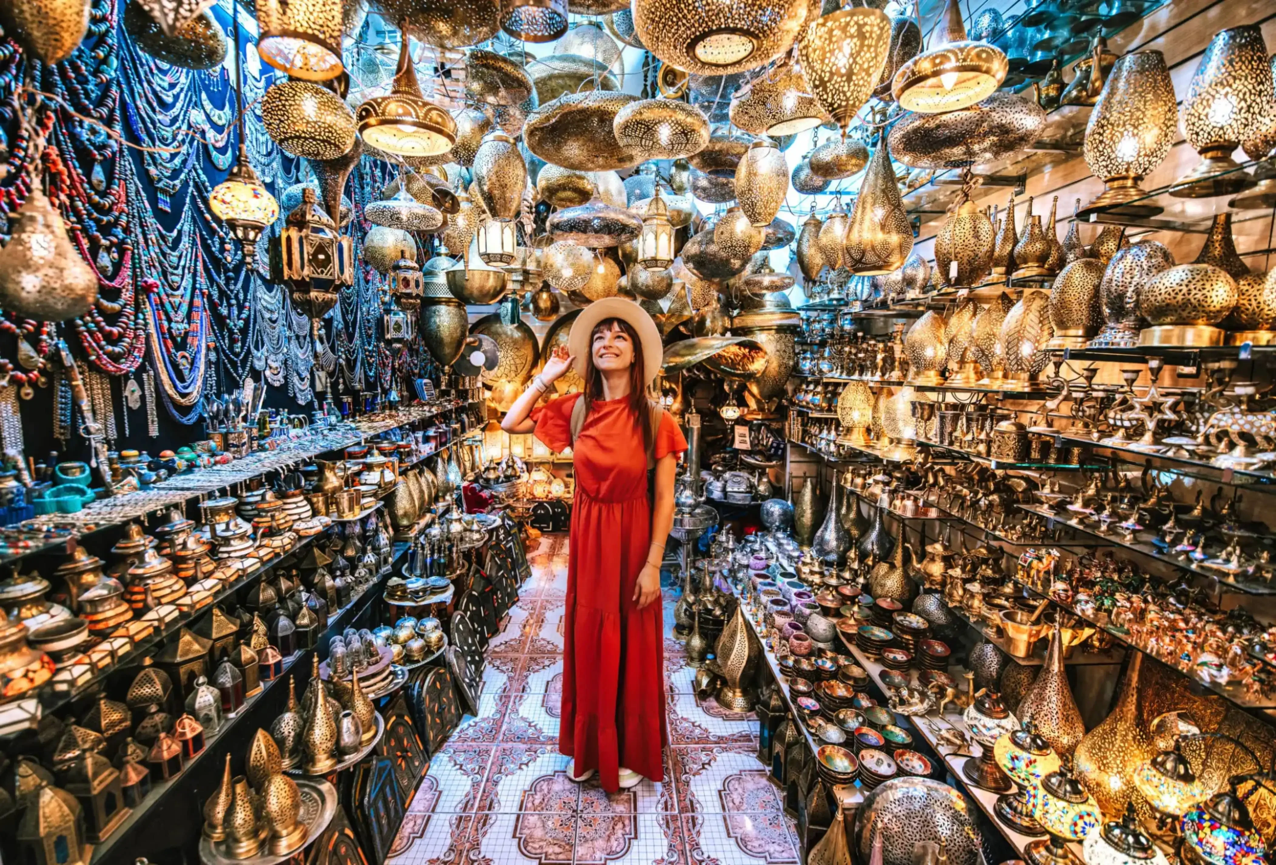 Smiling woman in red dress exploring a vibrant Middle Eastern or Moroccan-style lantern and brassware market filled with hanging lamps, trays, and colorful decor