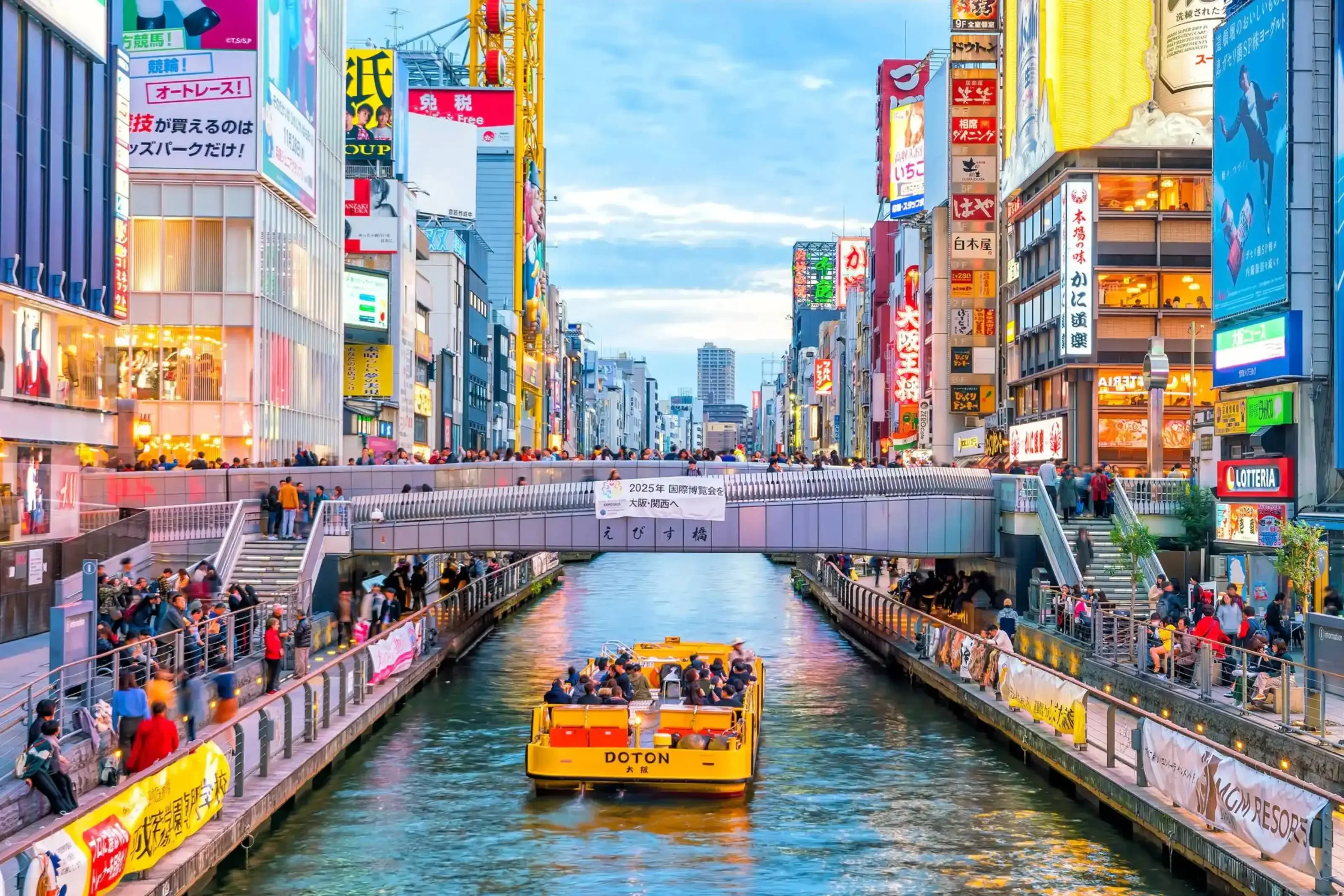 Tour boat cruising through the lively Dotonbori canal in Osaka, Japan, surrounded by neon billboards, busy walkways, and colorful storefronts