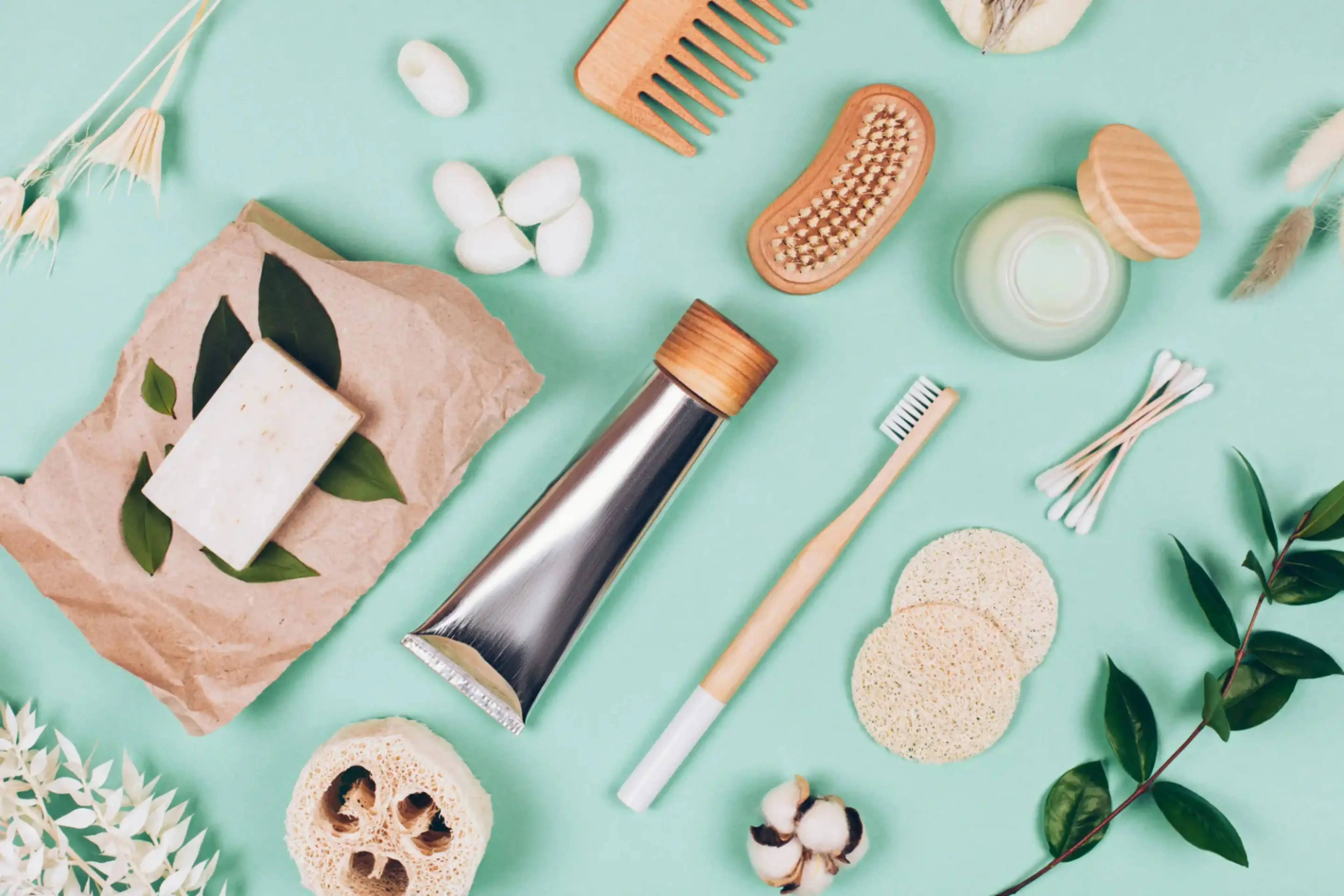 Flat lay of eco-friendly travel toiletries including bamboo toothbrush, wooden comb, reusable containers, and natural accessories on a pastel green background