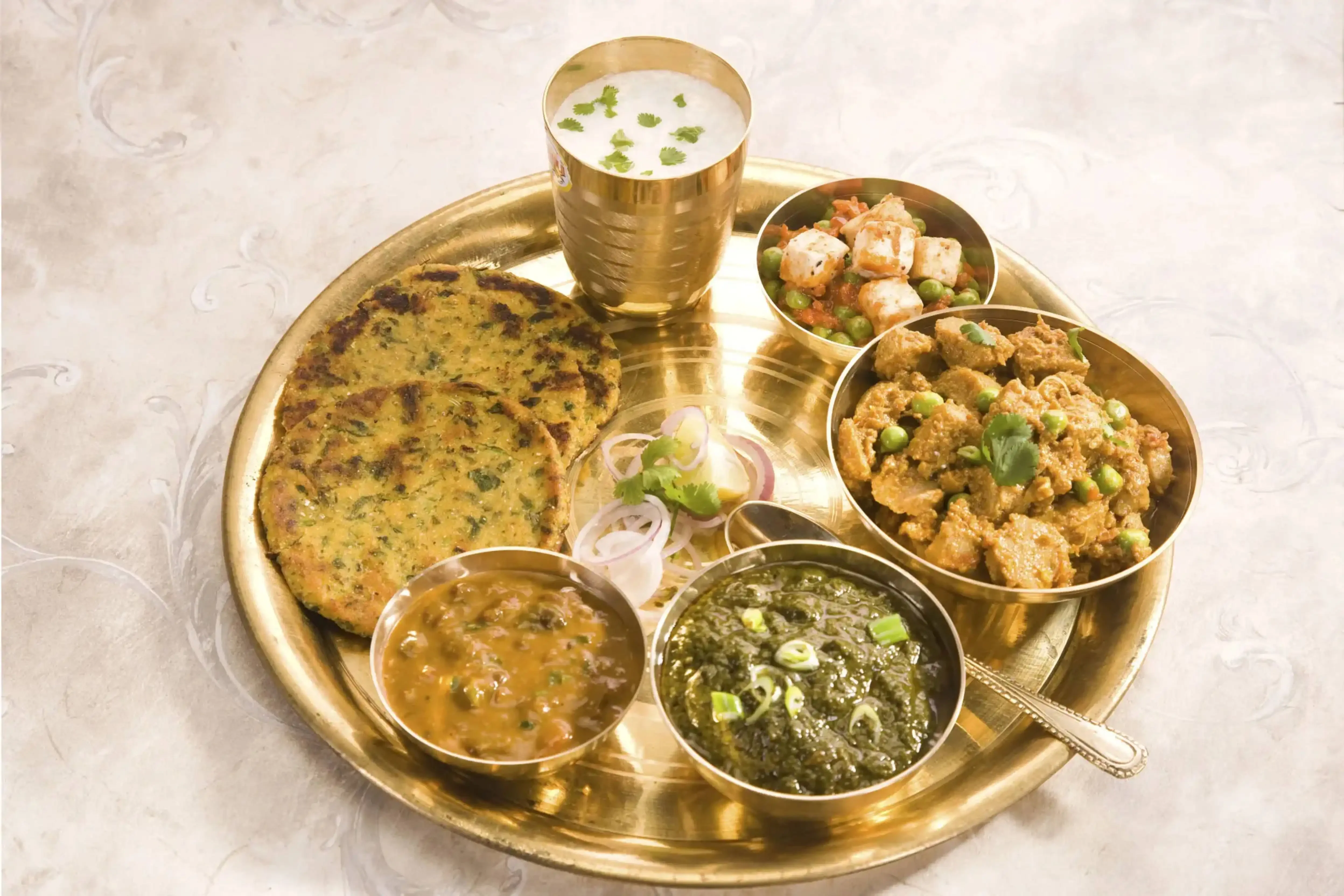 Authentic Indian thali with makki di roti, sarson da saag, mixed vegetables, lentils, and buttermilk served on a traditional brass plate