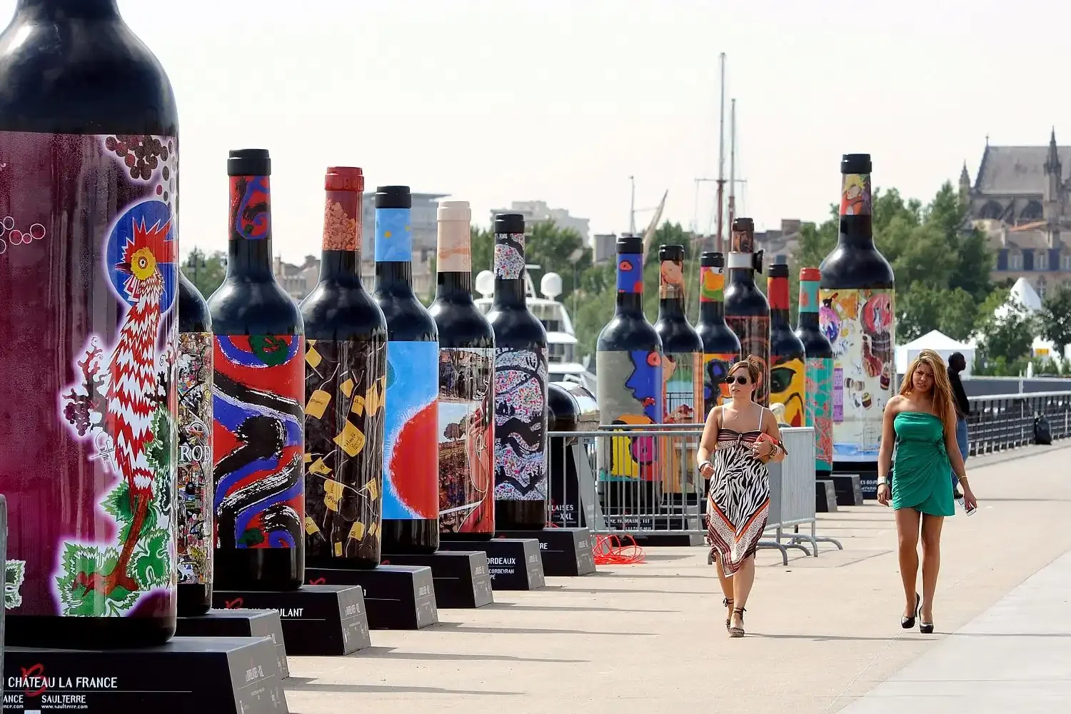 Outdoor art exhibition featuring giant wine bottles painted with colorful murals, with visitors walking along a waterfront promenade