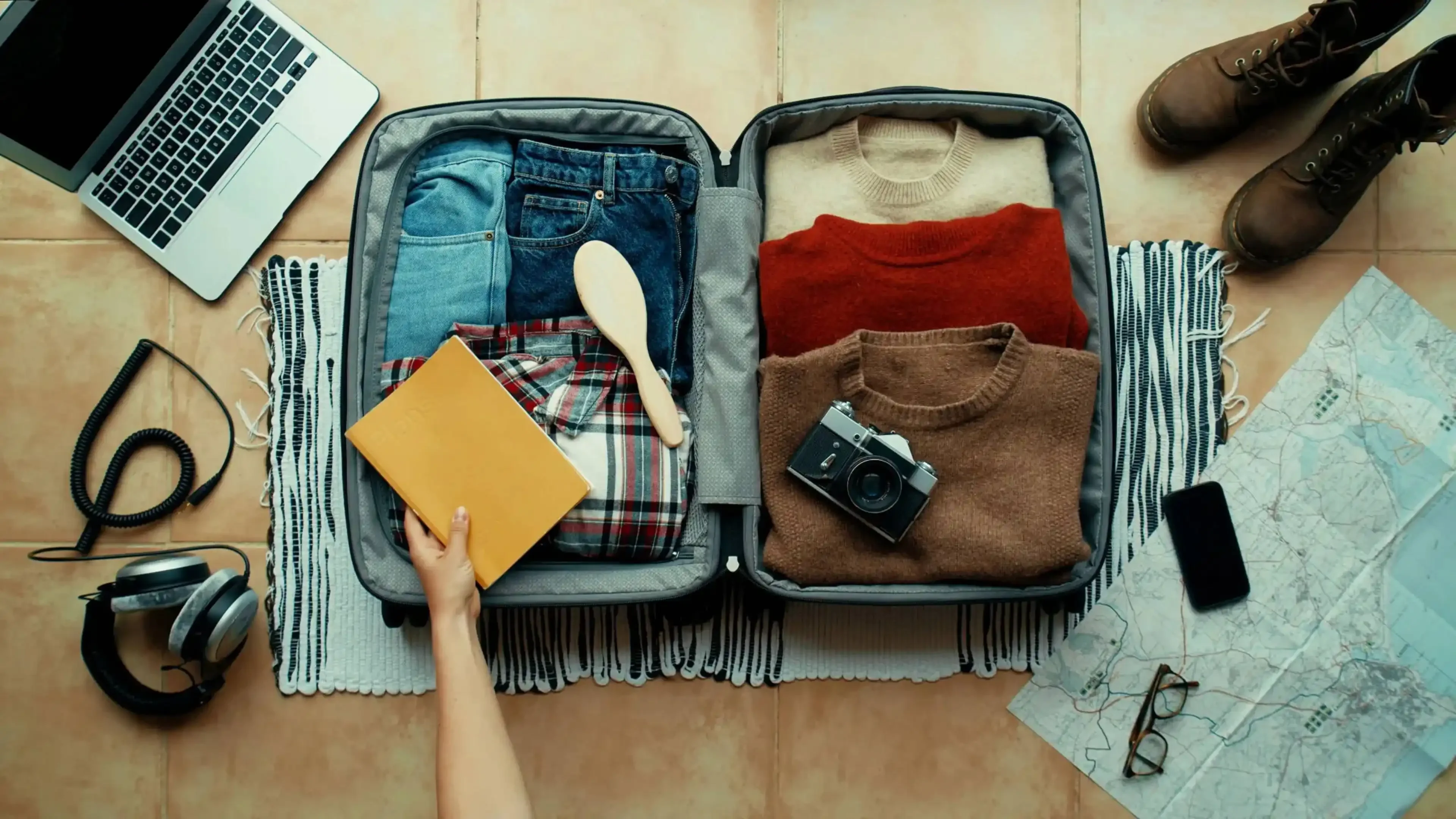 Top view of an open suitcase being packed with travel essentials including clothes, camera, and a travel journal