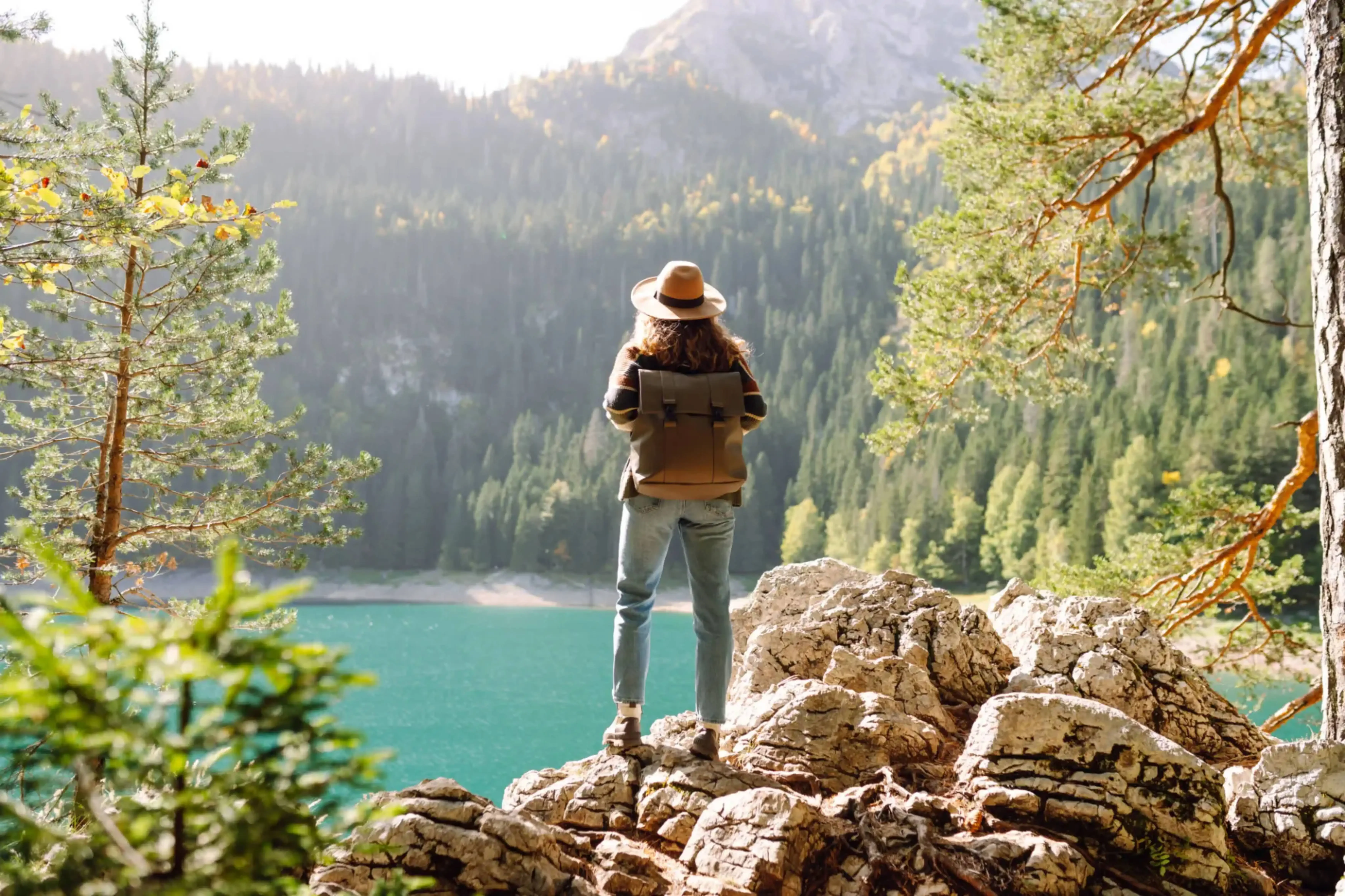 Female solo traveler with backpack and hat standing on rocky cliff overlooking turquoise alpine lake and forested mountains