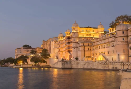 Evening view of Udaipur City Palace glowing with lights, reflecting in Lake Pichola, Rajasthan, India