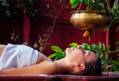 Woman receiving Ayurvedic Shirodhara therapy with oil stream on forehead in a serene outdoor spa setting