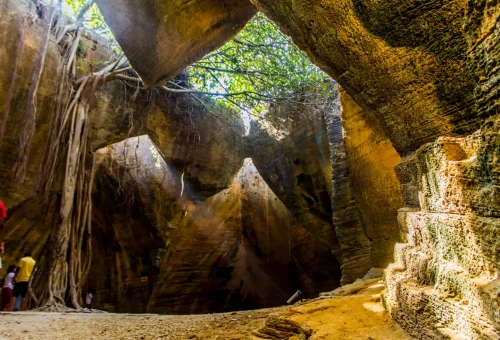 "Natural sandstone cave formations with tree roots and carved stone steps at Arwah Cave in Meghalaya, India"