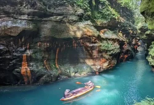 Wari_Chora-"Wooden boat gliding through crystal-clear turquoise river surrounded by lush cliffs and forest in Meghalaya, India for Assam Tours"