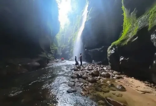 "Travelers walking through a narrow river canyon with sunlight streaming in and moss-covered rock walls in Sohra, Meghalaya"