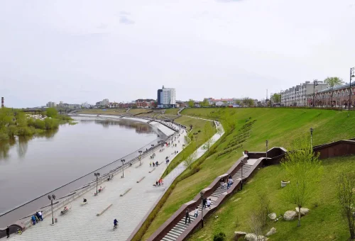 "Urban riverside park with walking paths, grassy slopes, and modern buildings in the background on a cloudy day"