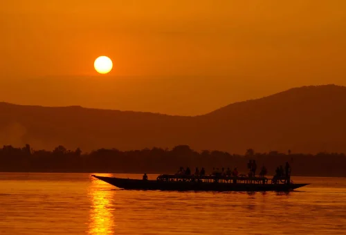 "Silhouetted boat gliding across a river at sunset with a glowing orange sky and distant hills in the background"