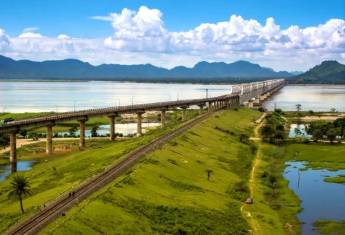 "Aerial view of a long bridge stretching over a river with green fields and distant mountains under a bright blue sky with clouds"
