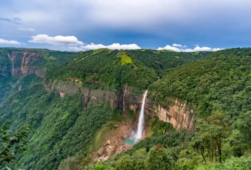 "Scenic view of a tall waterfall cascading down a lush green cliff into a turquoise pool surrounded by forested hills under a blue sky"